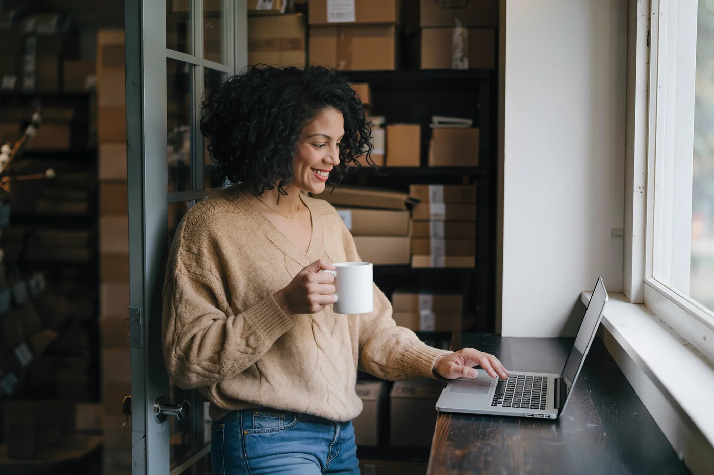 A shop owner with a mug of coffee at her laptop by the window of her stockroom, shelves of cardboard boxes behind her